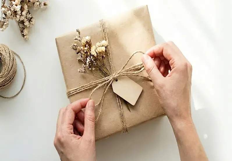 Manos atando un lazo en un regalo envuelto en papel kraft con flores secas y una etiqueta de madera con el logo de Cucurrina, destacando el cuidado en el detalle y el empaquetado artesanal.