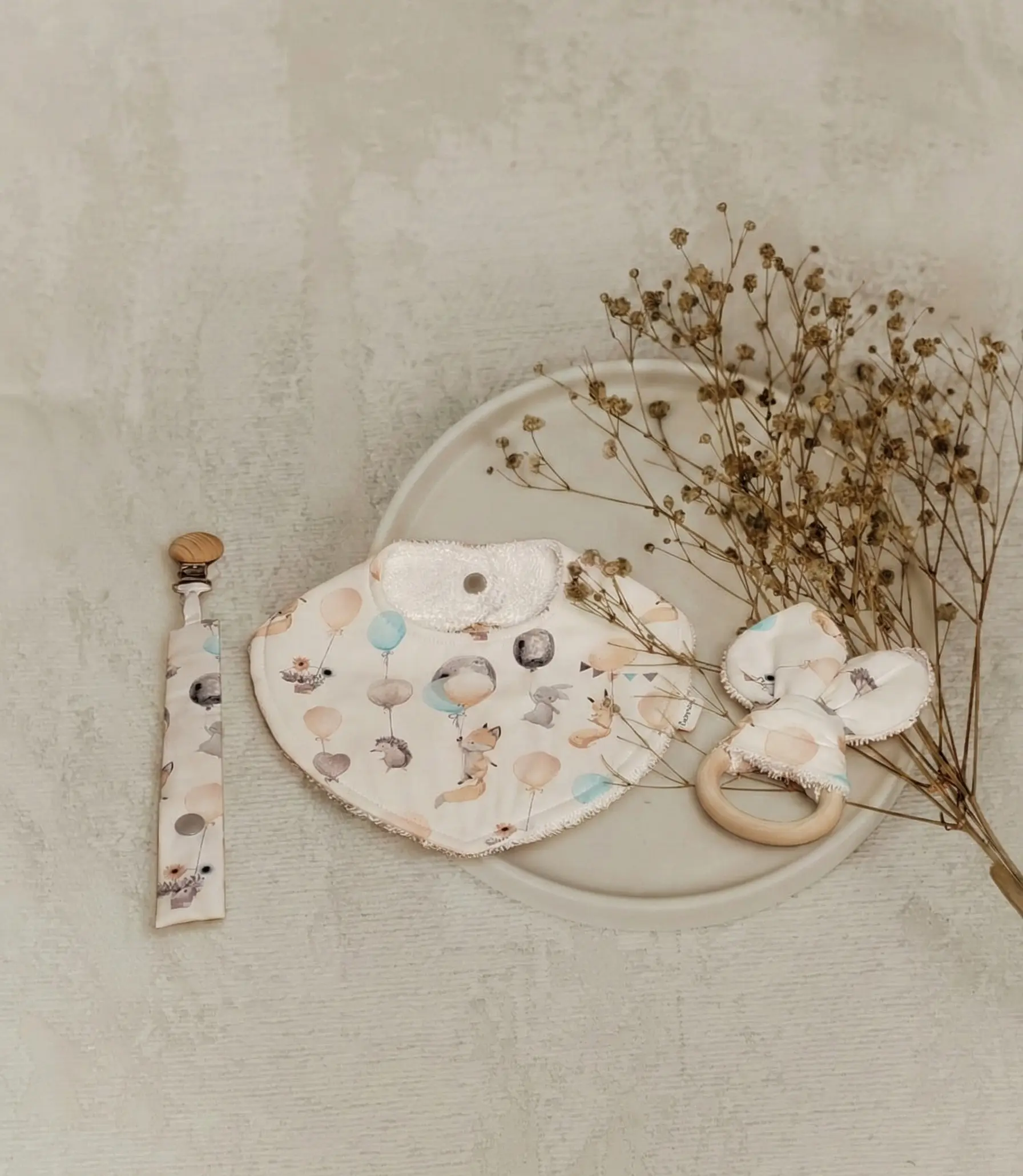 Composición de accesorios textiles para bebé hechos a mano en España: babero, chupetero y mordedor de orejas de conejo con estampado de animales y globos, marca Cucurrina.